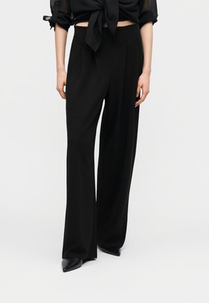 Kobieta w czarnych spodniach o wysokim stanie i szerokich nogawkach z plisami, w połączeniu z czarnymi spiczastymi butami i czarną przezroczystą bluzką wiązaną na kokardę.