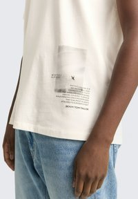 Witte katoenen t-shirt met een grafische print en tekst op de linkeronderzijde. In combinatie met lichtblauwe denim jeans, zichtbaar vanaf de taille naar beneden.