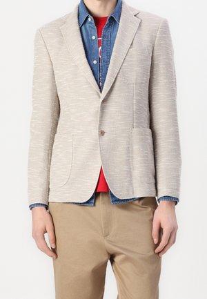 Persona que lleva una blazer beige texturizada sobre una camisa vaquera azul y una camiseta roja con pantalones beige, de pie frente a un fondo blanco.