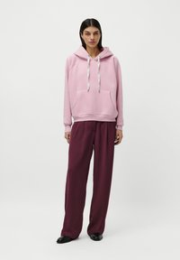 Femme se tenant debout, portant un sweat à capuche rose clair avec des cordons blancs, un pantalon large bourgogne et des chaussures noires sur un fond blanc uni.