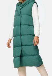 Giubbotto lungo imbottito verde con cappuccio, caratterizzato da un design trapuntato, chiusura a scatto e tasche laterali, abbinato a pantaloni aderenti beige.