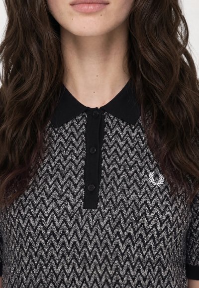 Femme aux longs cheveux ondulés et foncés portant un polo à motif zigzag noir et blanc avec un col et des boutons noirs.