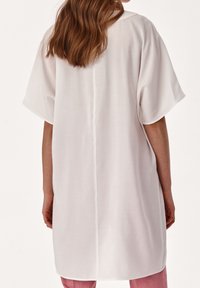 Donna con lunghi capelli rossastri indossa una camicia bianca lunga e ampia con maniche corte, abbinata a pantaloni rosa.