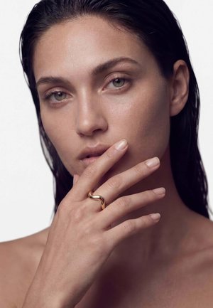 Femeie cu păr negru ud își atinge buzele cu degete subțiri purtând un inel auriu neted curbat pe degetul mijlociu, pe un fundal simplu.