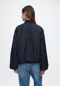 Tamsiai mėlynas bombonešio stiliaus švarkelis su lygiu paviršiumi, ribuotu apykakle ir elastingu apačios kraštu, dėvimas ant šviesiai mėlynų džinsų.