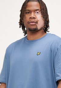 T-shirt en coton bleu clair avec un col rond, des manches courtes et un petit logo jaune brodé sur la poitrine. Texture lisse.
