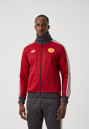 Rød lynlåsjakke med højt krave, hvide Adidas-striber og et Manchester United-logo på brystet. Lavet af glat stof. Grå bukser.