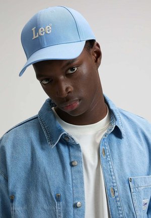 Junger Mann, der eine hellblaue Lee-Kappe und eine passende Jeansjacke über einem weißen T-Shirt trägt, schaut geradeaus mit neutralem Gesichtsausdruck.