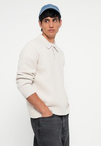 Light beige textured knit pullover with a collared neckline, paired with dark gray jeans, and a blue denim cap. Model stands with hands in pockets.