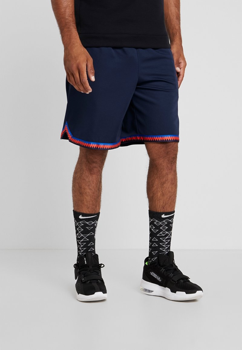 Homme portant un short de sport bleu marine avec une bordure rouge et bleue, des chaussettes Nike noires à motifs et des baskets Nike noires, debout sur un sol simple.