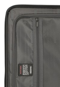 Interior de una bolsa de viaje gris con varios compartimentos, incluyendo un bolsillo de malla con cremallera y una etiqueta con información del producto.