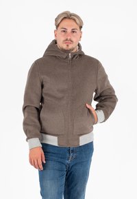 Uomo con capelli castano chiaro che indossa una giacca con cappuccio marrone con polsini grigi e jeans blu, in piedi con una mano in tasca.
