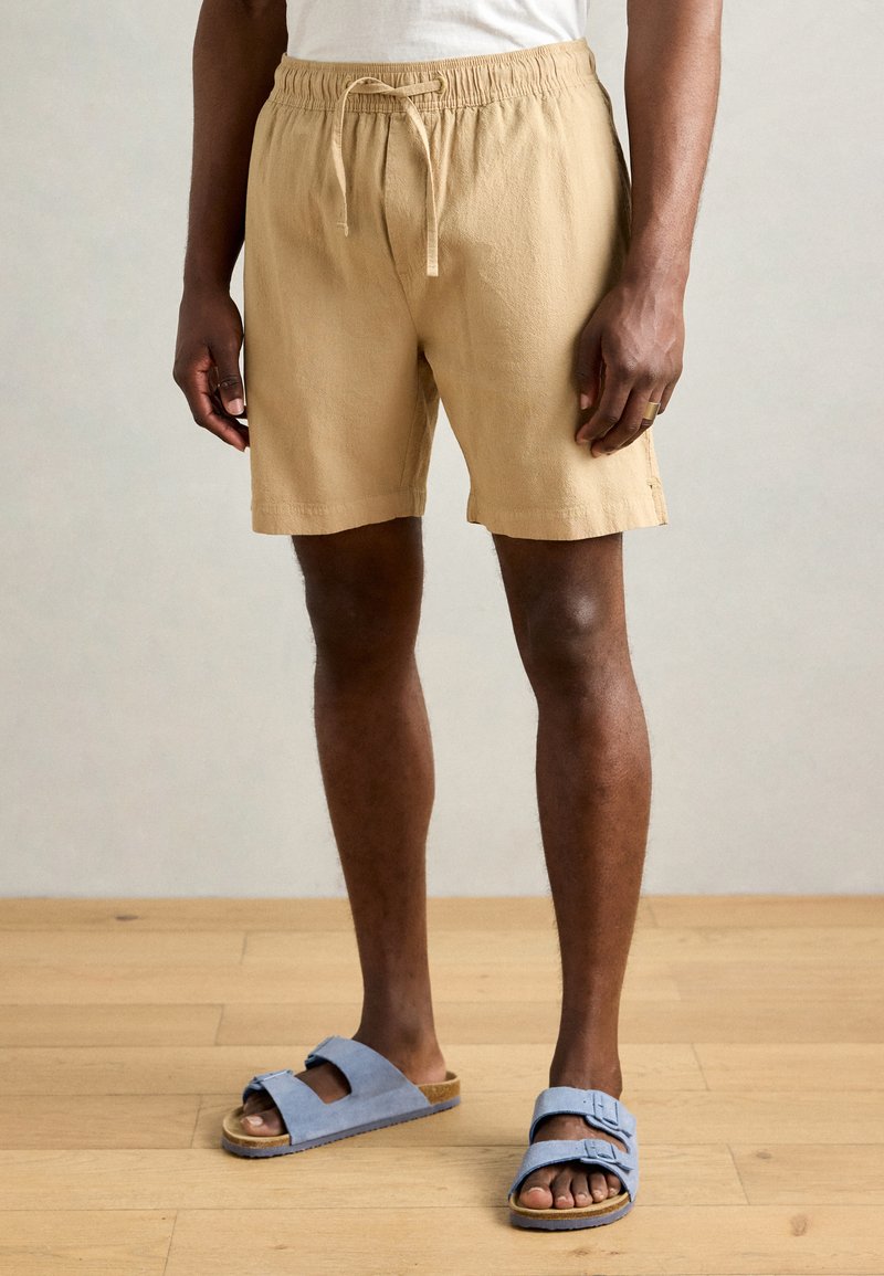 Khaki shorts med elastisk talje og snor. Lavet af tekstureret stof, har de sidelommer og en afslappet pasform. Sammen med blå sandaler.