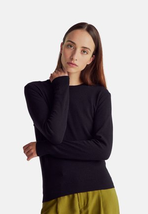Maglione nero a maniche lunghe con scollatura rotonda e polsini a coste. Texture morbida, design aderente, indossato con pantaloni verde oliva.
