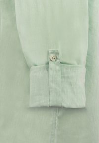 Chemise en lin vert clair avec des manchettes à bouton unique. Le tissu présente une surface légèrement texturée, d'apparence douce et légère.
