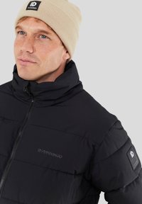 Doudoune noire avec col haut, fermeture éclair à l'avant et écusson logo sur la manche. Associée à un bonnet beige côtelé.