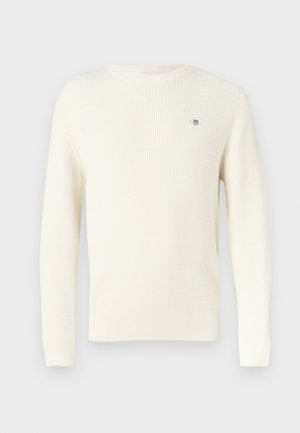 Kremowy sweter z fakturowanym wzorem dzianiny, ściągaczami na mankietach i dole oraz małym logo na piersi. Długie rękawy, okrągły dekolt.