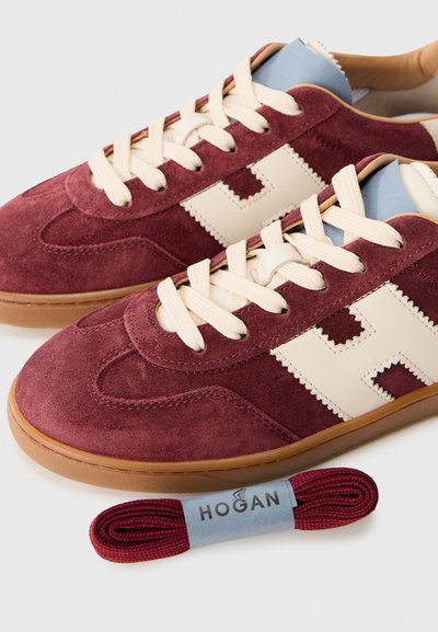 Un paio di sneakers in suede bordeaux con lacci beige, logo "H" beige sui lati, suole in gomma e un paio di lacci bordeaux di ricambio etichettati "HOGAN."