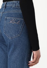 Denim jeans in medium blue with a fitted shape, featuring a back pocket accentuated by cursive embroidered text reading "Karl."