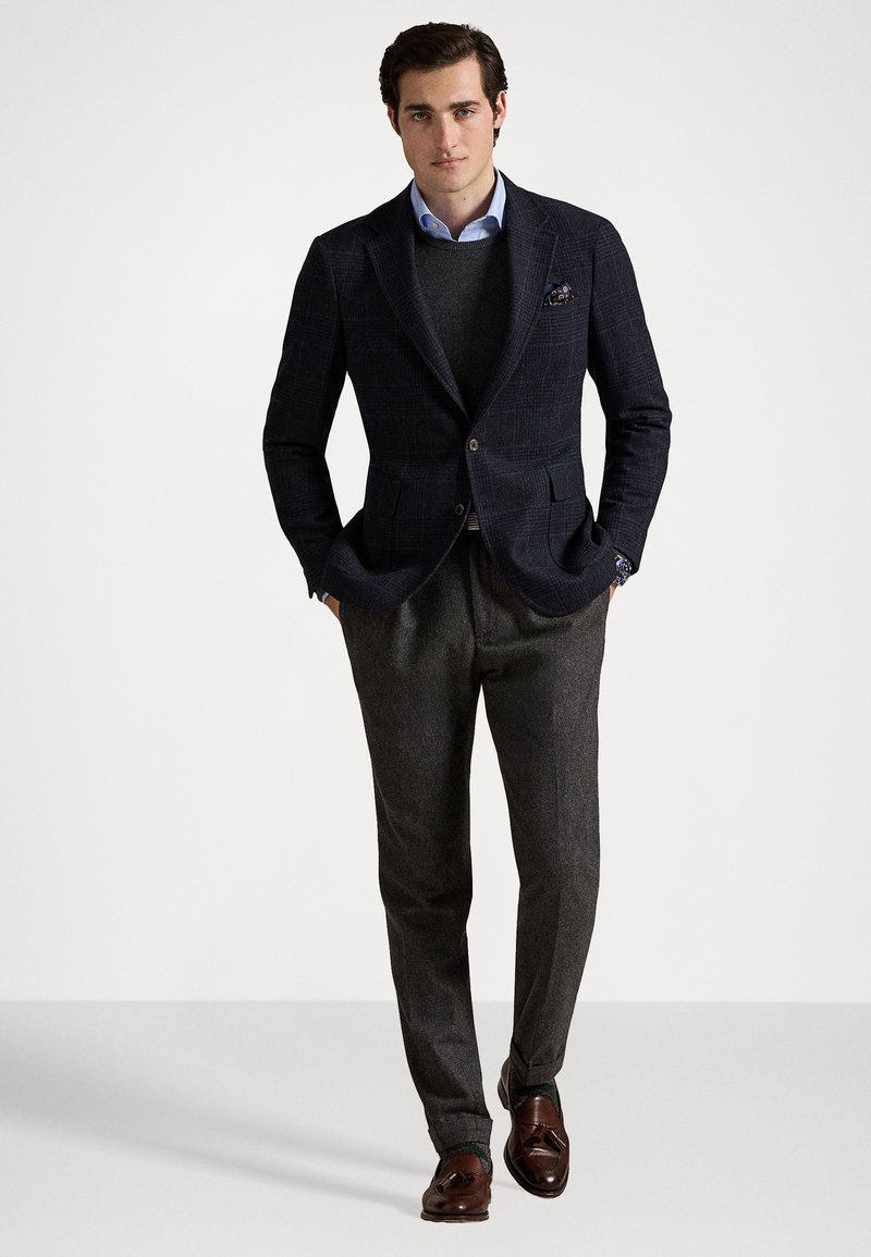Blazer a quadri navy, pullover grigio a maglia, camicia azzurra chiara. Pantaloni grigio scuro e mocassini marroni. Fazzoletto da taschino aggiunge un tocco di dettaglio. Silhouette a vestibilità classica.