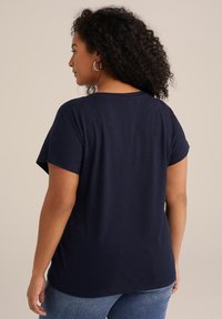 Kvinna med lockigt hår som bär en marinblå texturerad topp med korta ärmar och blå jeans, sedd bakifrån mot en enfärgad bakgrund.