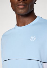 Camiseta de color azul claro con cuello redondo, que presenta una franja horizontal negra y un logo blanco en el pecho. Textura de tela suave.