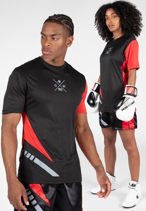 Zwarte sportshirt met rode mouwen, voorzien van een logo en geometrische accenten. Het mannelijke model draagt zwarte shorts met rode en grijze details.