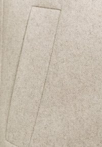Tejido de mezcla de lana color beige con una textura suave. Presenta un bolsillo tipo parche rectangular, líneas limpias y una superficie lisa y uniforme.