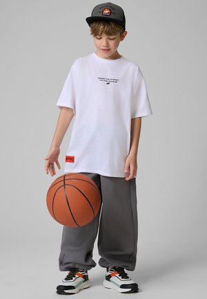 Młody chłopiec w czapce, za dużym białym t-shircie, szarych spodniach i trampkach odbijający piłkę do koszykówki na jednolitym szarym tle.