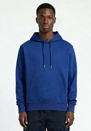 Giovane uomo in piedi, dritto, che indossa una felpa con cappuccio blu con tasca frontale e coulisse, abbinata a pantaloni neri su uno sfondo neutro.