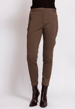 Woman wearing slim fit brown pants with zippered side pockets and dark brown heeled ankle boots, standing against a plain background.