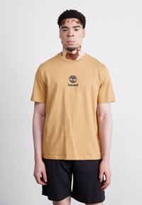 Sennepsgul bomulls T-skjorte med en svart Timberland-logo på brystet, sammen med svarte shorts. Enkelt, uten dekorasjoner.
