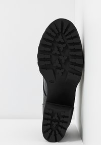 Semelle de botte noire avec des motifs de bande de roulement robustes, appuyée contre un mur blanc montrant un design détaillé pour l'adhérence.