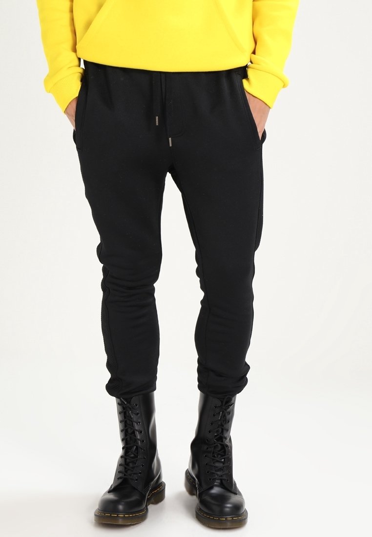 Černé zúžené tepláky s kapsami, stahovací šňůrkou v pase a texturovanými bočními panely, nošené s černými šněrovacími botami, na světlém pozadí.