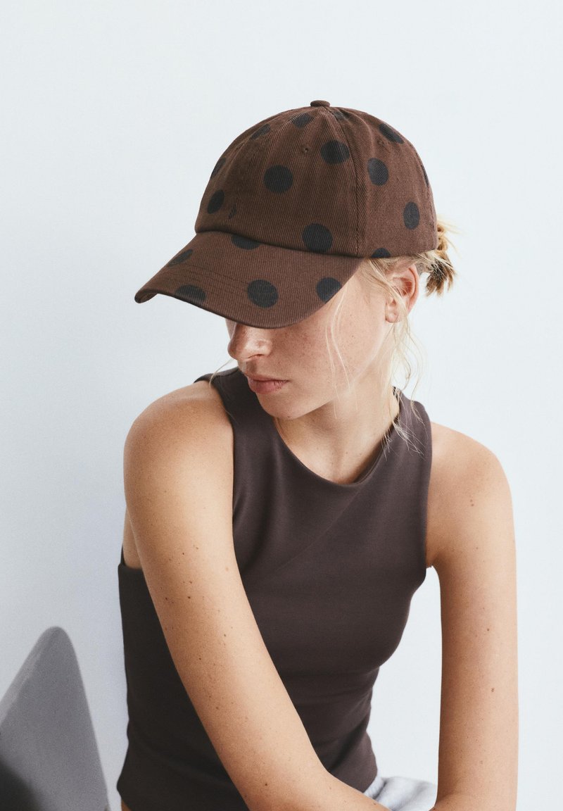Femme blonde portant un haut sans manches foncé et une casquette marron à pois noirs, regardant vers le bas et légèrement sur le côté.