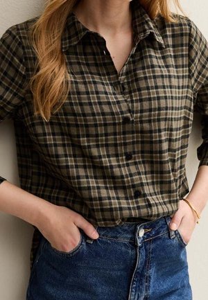 Vrouw draagt een groen-zwart geruit overhemd dat in een blauwe spijkerbroek is gestopt, met handen in de zakken.