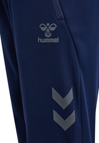 Marina atletiska byxor med en slät textur, med en logotyp på låret och två gråa chevron-accenter på sidan. Synlig ficka med dragkedja.