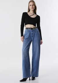 Haut court noir à côtes avec manches longues ; jeans large taille haute bleu clair avec une ceinture noire portant un texte blanc ; talons aiguilles noirs.