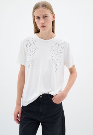 T-shirt blanc à manches courtes avec des clous en argent décoratifs en motif de feuilles sur la poitrine. Tissu doux, coupe décontractée, associé à un pantalon noir.