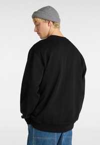 Felpa nera con collo a giro e polsini a costine, realizzata in tessuto morbido. Indossata con un berretto grigio piegato e jeans blu. Design semplice.