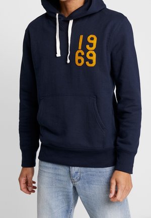 Homme portant un sweat à capuche bleu marine avec des cordons blancs et un patch jaune moutarde "1969", associé à un jean bleu clair.