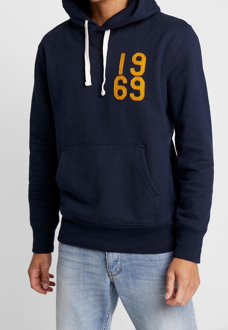 Homme portant un sweat à capuche bleu marine avec des cordons blancs et un patch jaune moutarde "1969", associé à un jean bleu clair.