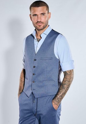 Mann mit kurzen Haaren und Tattoos, der ein hellblaues Hemd mit hochgekrempelten Ärmeln sowie eine blaue, geknöpfte Weste und Hose trägt und die Hände in den Taschen hat.
