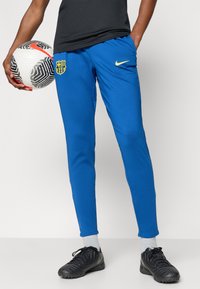 Μπλε αθλητικά παντελόνια από ελαφρύ υλικό, με λογότυπο της Μπαρσελόνα και κίτρινο σήμα της Nike, συνδυασμένα με μαύρα ποδοσφαιρικά παπούτσια.