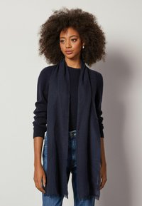 Mørkeblå tørklæde lavet af et blødt, tekstureret stof med en frynset kant. Båret over en mørk sweater, tilføjer det varme til afslappet påklædning.