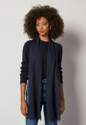 Mørkeblå tørklæde lavet af et blødt, tekstureret stof med en frynset kant. Båret over en mørk sweater, tilføjer det varme til afslappet påklædning.