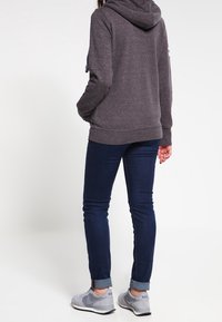 Sudadera gris oscuro con textura moteada, con capucha y bolsillos frontales, combinada con jeans azul oscuro y zapatillas gris.