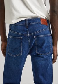 Blå denimjeans med rak passform, med två bakfickor med ett distinkt sömnadsmönster och en brun läderlogotyp.