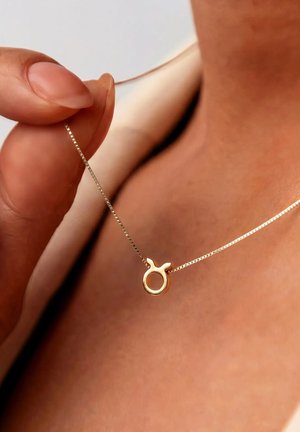 Hand, die eine zarte Goldkette mit einem kleinen, runden Anhänger hält, der zwei nach oben gerichtete Hörner zeigt, vor Haut und beiger Kleidung.