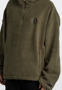 Sudadera de forro polar verde oliva con cuello de media cremallera, un logo circular, bolsillos laterales y tejido texturizado para mayor calor. Es de corte amplio y casual.